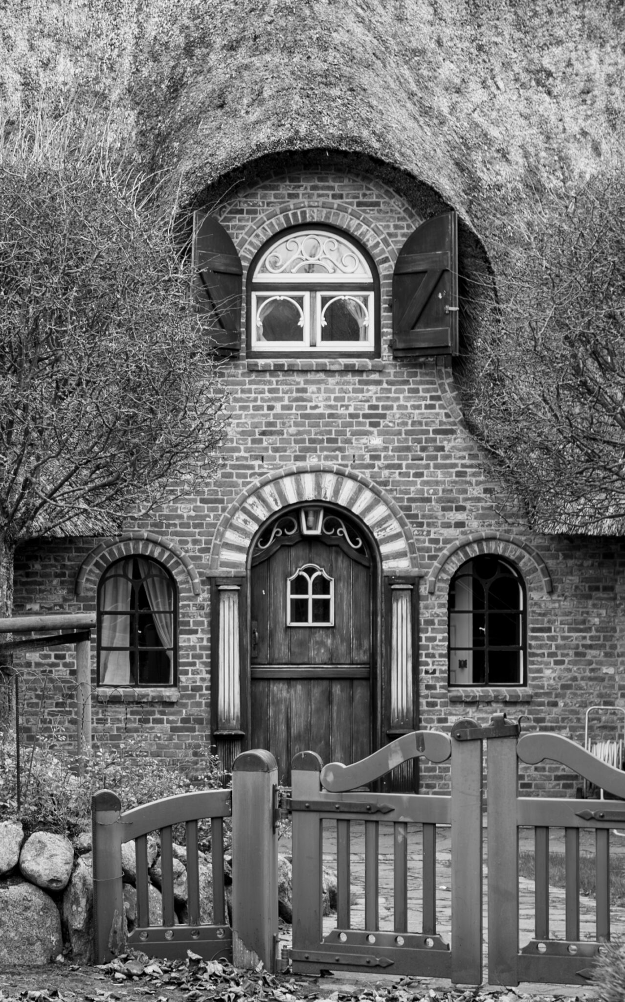 Ein Reet-gedecktes Haus in schwarz-weiss. Zusehen ist ein Teil des Hauses mit dem Eingangsbereich. Im Vordergrund sieht man einen Teil des Gartentors, die Eingangstür und drei Rundbogenfenster. Es ist ein für Norddeutschland typisches Reet- und Klinkerhaus.